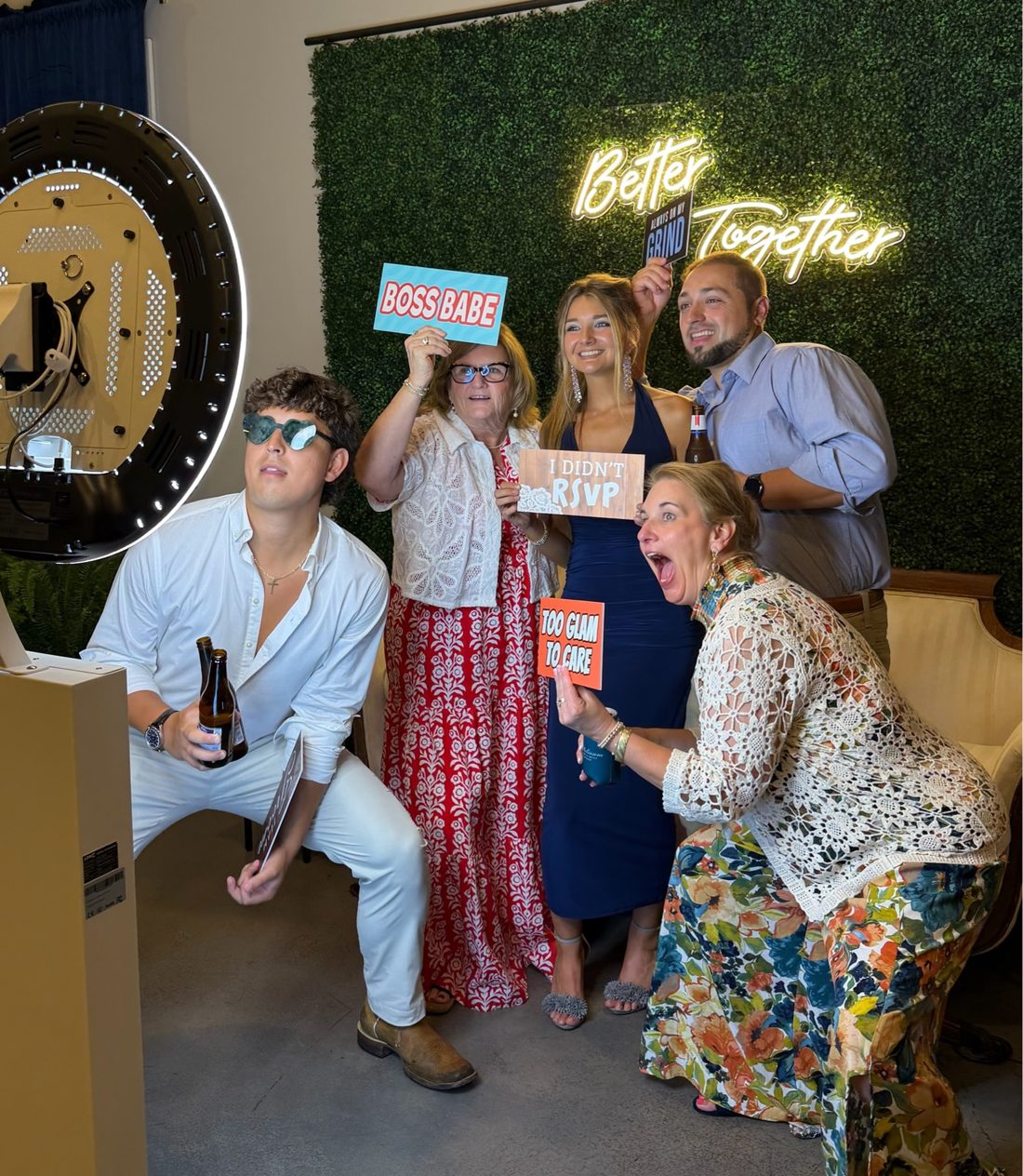 Wedding guests laughing with prop signs in front of the 'Better Together' neon greenery wall beside the photo booth